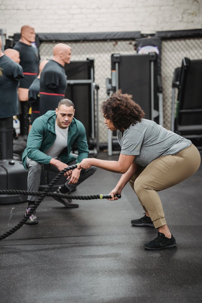 Full body focused plump African American female in activewear exercising with battle ropes near professional male coach in modern sports center