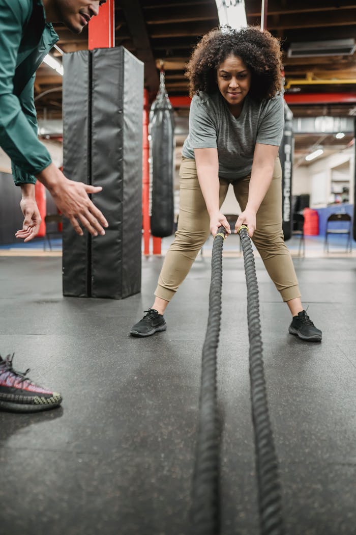 Positive plus sized African American female in sportswear with battle ropes listening to professional male trainer instructions in gym