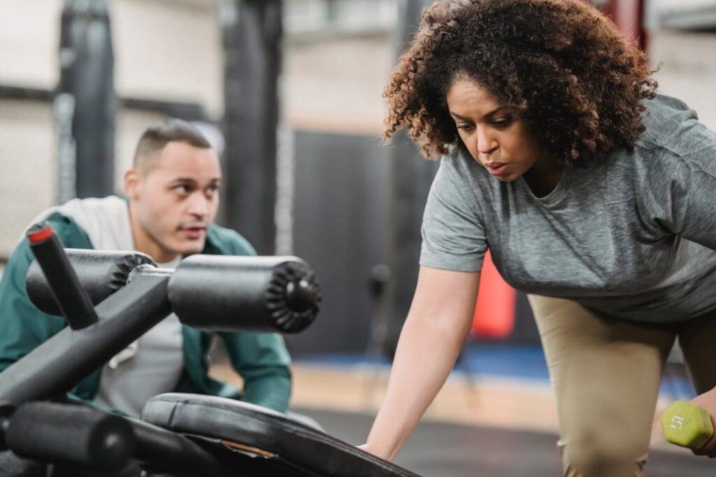 Experienced coach controlling crop African American female working out with dumbbell on blurred background of gym